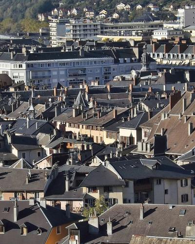 A high-angle view of a dense European cityscape featuring numerous slate and tile rooftops, chimneys, and historic buildings, with a modern white building and a hillside in the background.