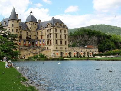 A large Renaissance-style château with conical towers and a domed roof, situated beside a lake with people relaxing on the grassy bank and French flags hanging from the facade.