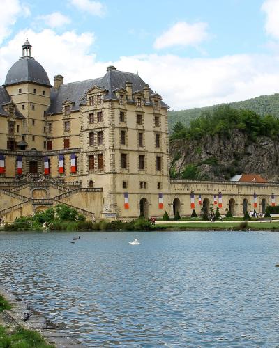 A large Renaissance-style château with conical towers and a domed roof, situated beside a lake with people relaxing on the grassy bank and French flags hanging from the facade.