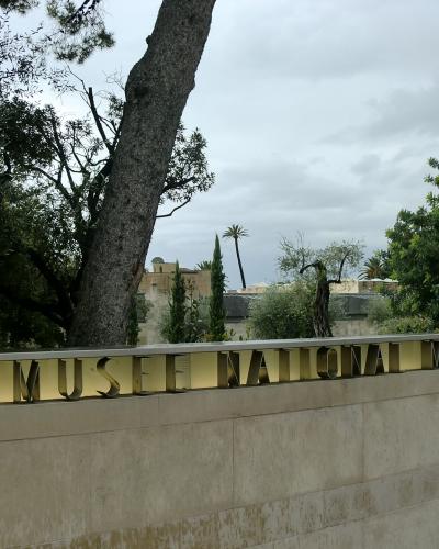 A low stone wall with large, gold-colored metal letters spelling "MUSEE NATIONAL MARC CHAGALL" set against a backdrop of trees and a cloudy sky.