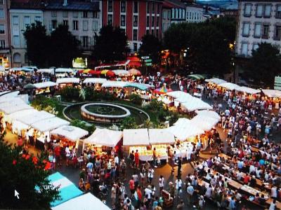 An elevated view of a crowded outdoor market in a European town square at dusk, featuring a central circular fountain and garden surrounded by rows of white market stalls and long communal dining tables filled with people.