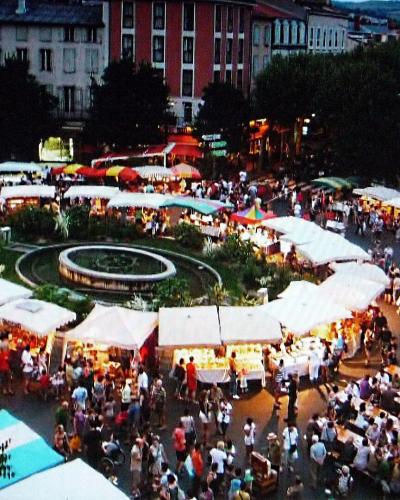 An elevated view of a crowded outdoor market in a European town square at dusk, featuring a central circular fountain and garden surrounded by rows of white market stalls and long communal dining tables filled with people.