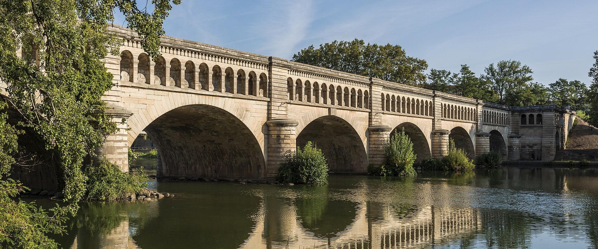 A stone aqueduct bridge with multiple large arches spanning a calm river, featuring a row of smaller arched openings along its upper deck, surrounded by green trees under a blue sky.