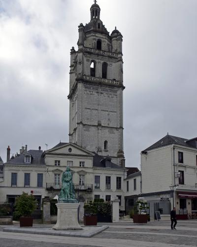 A tall, ornate stone bell tower (the Tour de l'Horloge) rises behind a statue of Alfred de Vigny in a paved town square in Loches, France.
