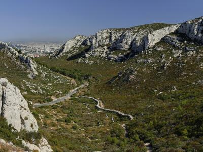 A panoramic view of a rocky, Mediterranean-style mountain pass with a winding road and hiking trails, overlooking a distant urban area under a clear blue sky.
