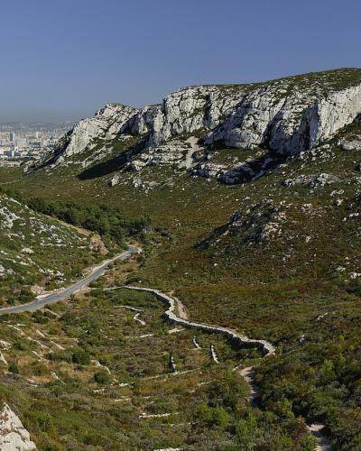 A panoramic view of a rocky, Mediterranean-style mountain pass with a winding road and hiking trails, overlooking a distant urban area under a clear blue sky.
