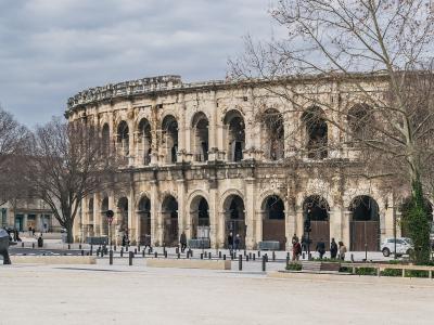 A large, two-tiered Roman amphitheater with arched openings, situated in a paved public square with bare trees and pedestrians.