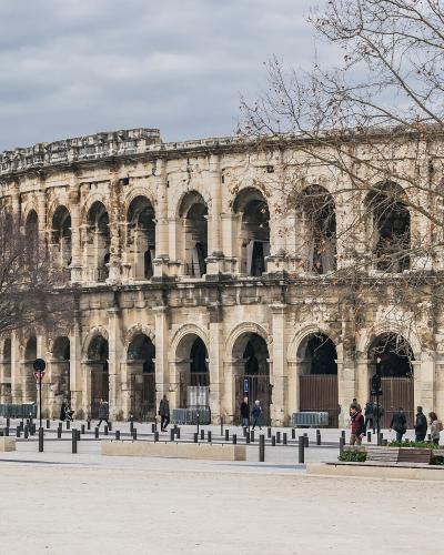 A large, two-tiered Roman amphitheater with arched openings, situated in a paved public square with bare trees and pedestrians.