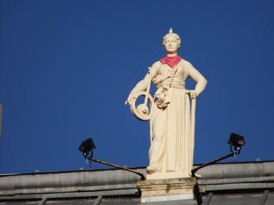 A close-up of a white neoclassical statue of a female figure wearing a red scarf, holding a lyre and staff, mounted on a pedestal atop a building roof against a clear blue sky, with two black floodlights visible below.