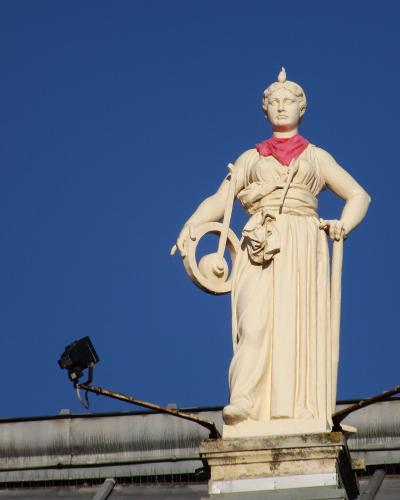 A close-up of a white neoclassical statue of a female figure wearing a red scarf, holding a lyre and staff, mounted on a pedestal atop a building roof against a clear blue sky, with two black floodlights visible below.