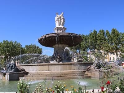 A large, multi-tiered stone fountain with sculpted figures including lions at the base and two standing statues atop a central basin, set in an urban plaza with trees and buildings in the background under a clear blue sky.