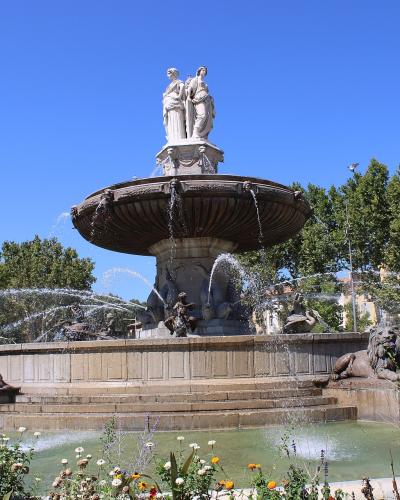 A large, multi-tiered stone fountain with sculpted figures including lions at the base and two standing statues atop a central basin, set in an urban plaza with trees and buildings in the background under a clear blue sky.