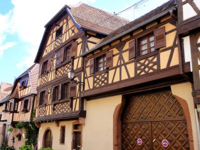 A low-angle view of traditional Alsatian half-timbered buildings with yellow stucco, dark timber framing, and a large wooden gate featuring diamond-patterned panels and two circular blue-and-red traffic signs.