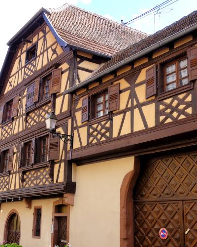 A low-angle view of traditional Alsatian half-timbered buildings with yellow stucco, dark timber framing, and a large wooden gate featuring diamond-patterned panels and two circular blue-and-red traffic signs.