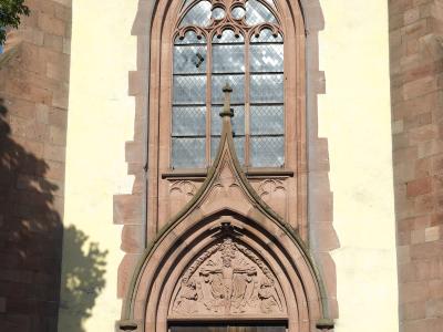 The image shows a close-up of a Gothic-style church entrance featuring a pointed arch doorway with a sculpted tympanum, a tall tracery window above it, and reddish sandstone framing against a pale yellow stucco wall.