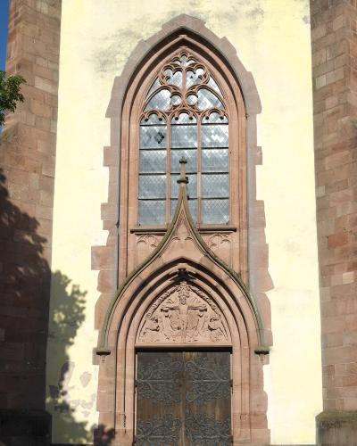 The image shows a close-up of a Gothic-style church entrance featuring a pointed arch doorway with a sculpted tympanum, a tall tracery window above it, and reddish sandstone framing against a pale yellow stucco wall.