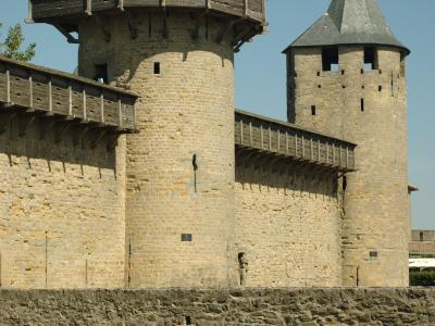 The image shows two stone towers with conical slate roofs and wooden hoardings, connected by a stone wall and a stone arch bridge in the foreground, under a clear blue sky.
