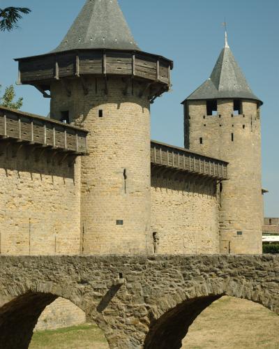 The image shows two stone towers with conical slate roofs and wooden hoardings, connected by a stone wall and a stone arch bridge in the foreground, under a clear blue sky.