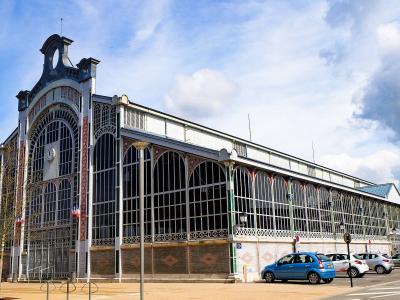 A large, ornate iron-and-glass market hall building with a prominent arched entrance, decorative brickwork, and French flags hanging from the facade, situated next to a parking lot with cars under a blue sky.