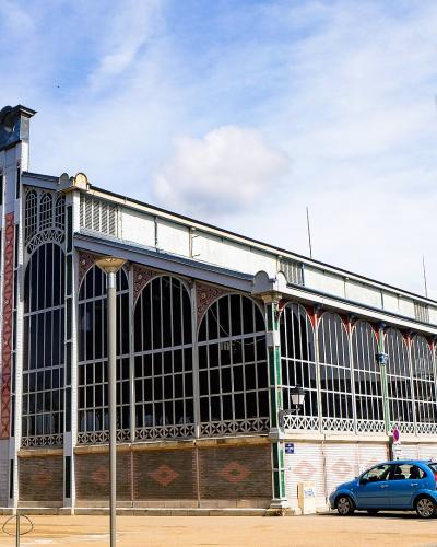 A large, ornate iron-and-glass market hall building with a prominent arched entrance, decorative brickwork, and French flags hanging from the facade, situated next to a parking lot with cars under a blue sky.