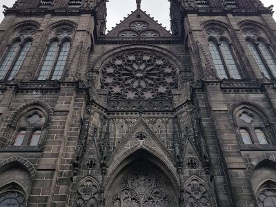A low-angle photograph of the dark stone Gothic facade of a cathedral featuring two prominent spires, a large rose window, and an ornate central portal with red doors.