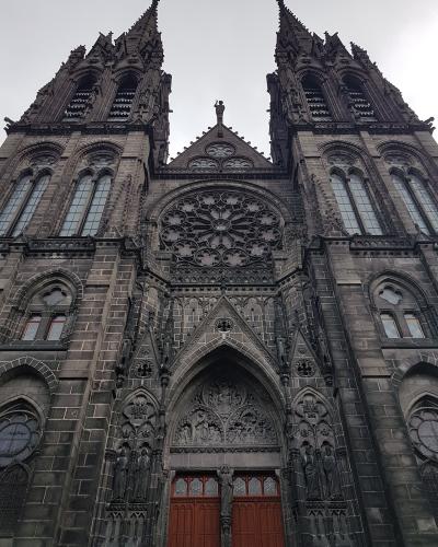 A low-angle photograph of the dark stone Gothic facade of a cathedral featuring two prominent spires, a large rose window, and an ornate central portal with red doors.