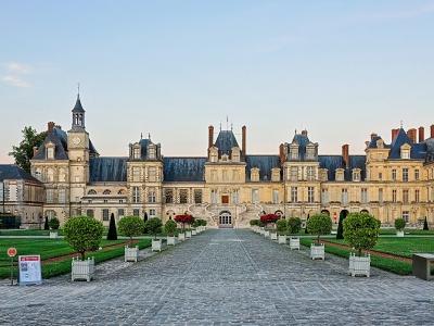 A wide-angle photograph of the Cour d'Honneur (Courtyard of Honor) at the Château de Fontainebleau, showing the central pavilion with its clock tower, flanked by long wings of the palace, and a formal garden with manicured lawns, topiary, and a co