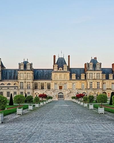 A wide-angle photograph of the Cour d'Honneur (Courtyard of Honor) at the Château de Fontainebleau, showing the central pavilion with its clock tower, flanked by long wings of the palace, and a formal garden with manicured lawns, topiary, and a co