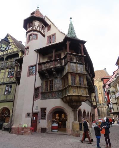 A historic half-timbered and stucco building with ornate wooden balconies, painted murals on its façade, a conical green roof turret, and a corner tower with a red-tiled roof; people are walking on a cobblestone street in front, and signage includ