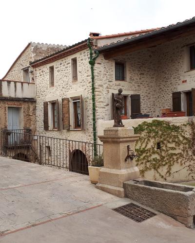 The image shows a stone-paved village square with traditional stone buildings, a central stone fountain topped by a bronze statue, and a black metal railing along the edge of a drop-off.