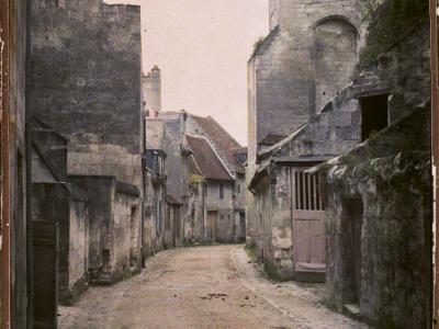 A vintage colorized photograph of a narrow, unpaved medieval street flanked by old stone buildings and a large stone tower or fortification on the right.