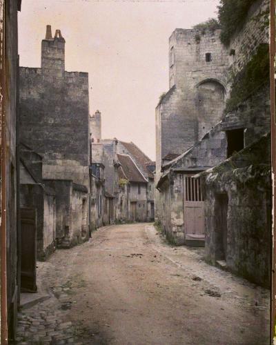 A vintage colorized photograph of a narrow, unpaved medieval street flanked by old stone buildings and a large stone tower or fortification on the right.