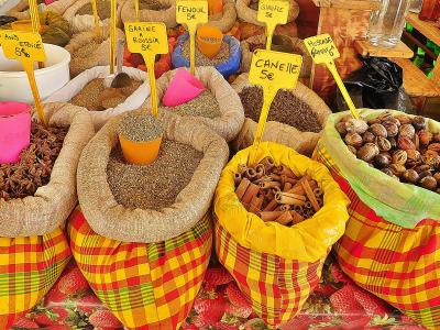 The image shows a close-up of an open-air market stall displaying bulk spices like star anise, cinnamon, and fenugreek in colorful checkered sacks with yellow price signs written in French (e.g., "ANIS ETOILE 5€", "CANNELLE 5€").