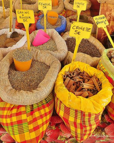 The image shows a close-up of an open-air market stall displaying bulk spices like star anise, cinnamon, and fenugreek in colorful checkered sacks with yellow price signs written in French (e.g., "ANIS ETOILE 5€", "CANNELLE 5€").