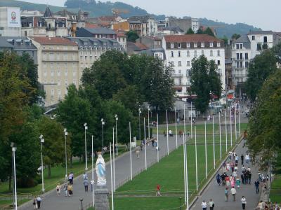 A wide pedestrian promenade lined with white flagpoles and flanked by green lawns, featuring a prominent statue of the Virgin Mary on a stone pedestal in the foreground, with multi-story buildings and hills in the background; signage for “SALON IN