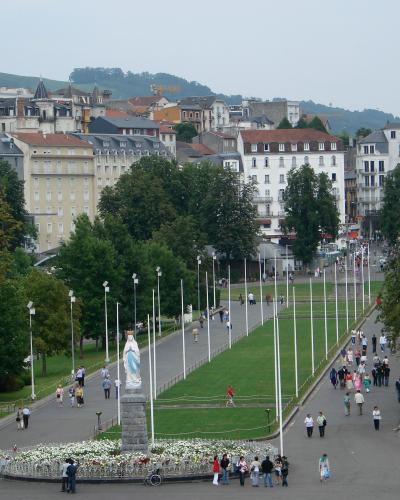 A wide pedestrian promenade lined with white flagpoles and flanked by green lawns, featuring a prominent statue of the Virgin Mary on a stone pedestal in the foreground, with multi-story buildings and hills in the background; signage for “SALON IN