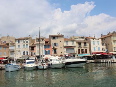 A view of a harbor lined with multi-story buildings featuring shuttered windows and terracotta roofs, with several boats moored in the foreground under a partly cloudy sky.