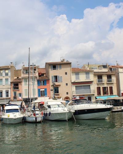 A view of a harbor lined with multi-story buildings featuring shuttered windows and terracotta roofs, with several boats moored in the foreground under a partly cloudy sky.