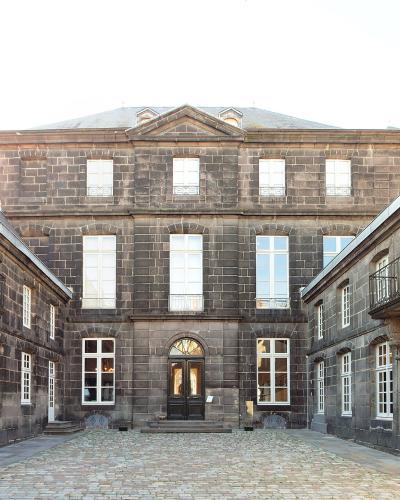 A courtyard view of a historic stone mansion with dark masonry, white-framed windows, and a central entrance, flanked by wings with white garage-style doors and iron balconies.