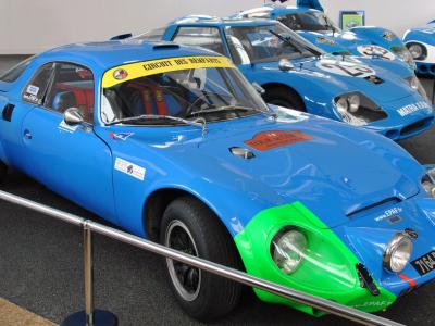A blue Matra 610 "Coupé Napoléon" race car with a green front fender, displayed indoors behind a stanchion rope, with other blue race cars visible in the background.