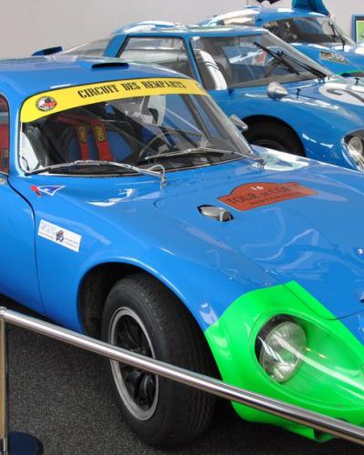 A blue Matra 610 "Coupé Napoléon" race car with a green front fender, displayed indoors behind a stanchion rope, with other blue race cars visible in the background.