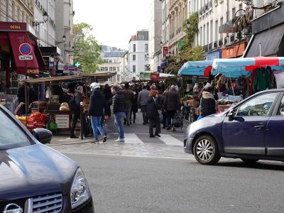 Bustling outdoor market stalls line Rue d'Aligre in Paris with shoppers and cars in the foreground.