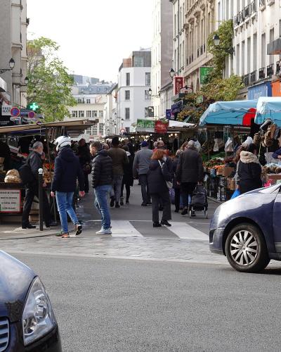 Bustling outdoor market stalls line Rue d'Aligre in Paris with shoppers and cars in the foreground.