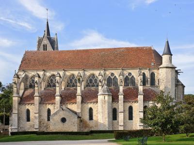 The image shows the exterior of a large Gothic-style stone church with a long nave, flying buttresses, a central spire, and a smaller turret on the right, set against a blue sky with some clouds and greenery in the foreground.