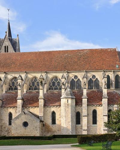 The image shows the exterior of a large Gothic-style stone church with a long nave, flying buttresses, a central spire, and a smaller turret on the right, set against a blue sky with some clouds and greenery in the foreground.