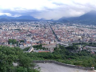 A panoramic view of Grenoble, France, showing the cityscape with red-tiled roofs, surrounded by green hills and mountains under a cloudy sky, with part of a cable car structure visible on the left.