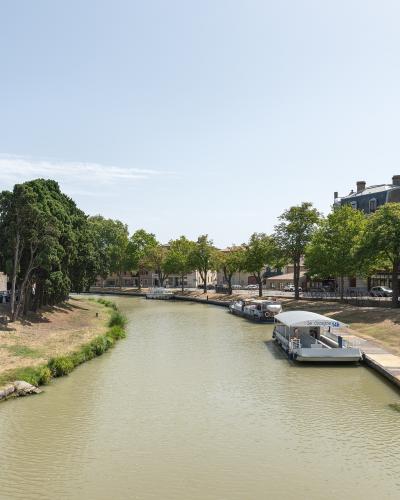 A wide, calm canal with tour boats docked along the right bank, lined with green trees and buildings under a clear sky; one boat displays "Le Cocagne 4h".