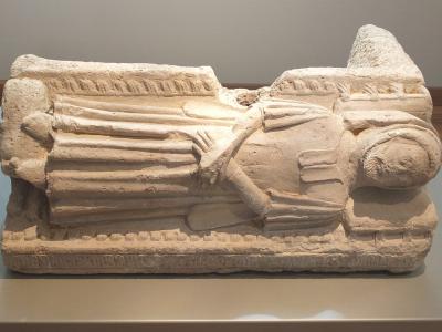 A stone sarcophagus lid featuring a carved effigy of a robed figure with hands clasped in prayer, displayed on a plinth in a museum gallery setting.