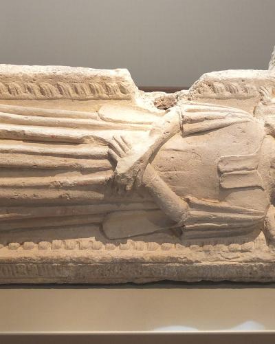 A stone sarcophagus lid featuring a carved effigy of a robed figure with hands clasped in prayer, displayed on a plinth in a museum gallery setting.