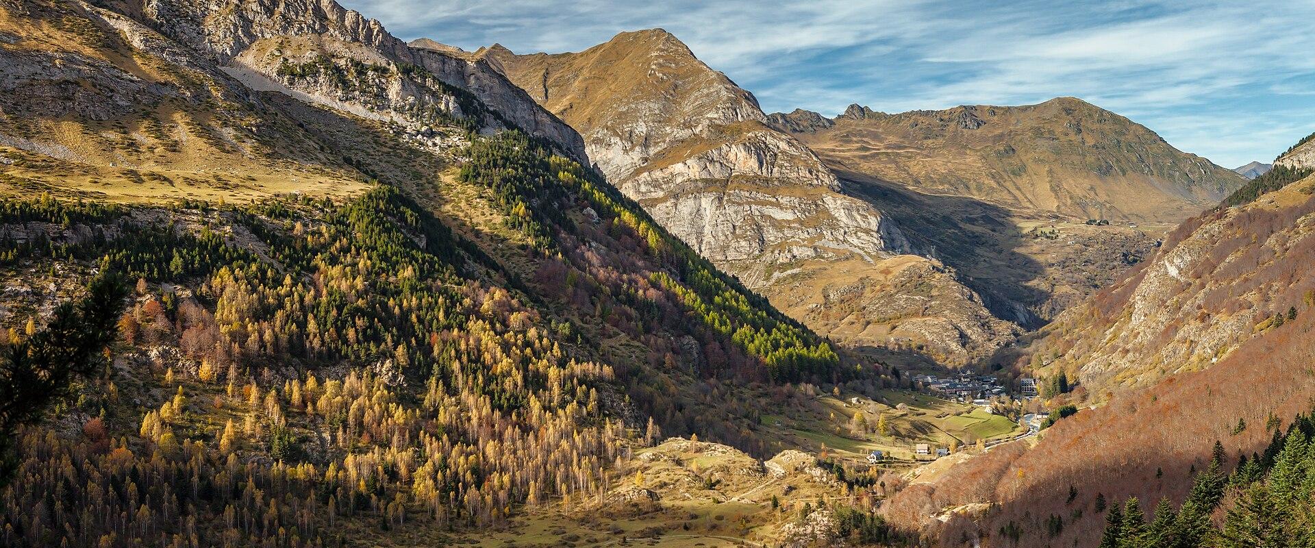 A high-angle landscape photograph of a mountain valley with autumn-colored larch trees, coniferous forests, rocky slopes, and a small village nestled in the distance under a partly cloudy sky.